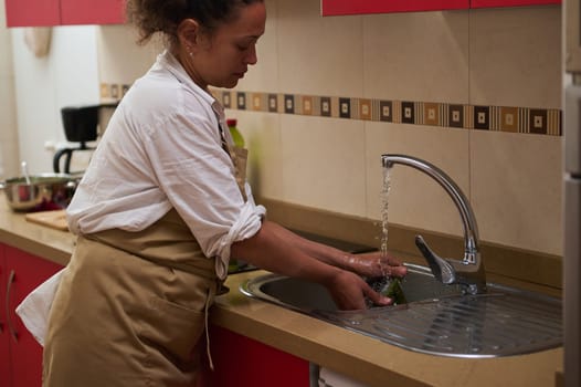 A woman in an apron is washing fresh vegetables under running water in a modern kitchen. The scene emphasizes hygiene and healthy cooking practices, highlighting a cozy, everyday domestic life.