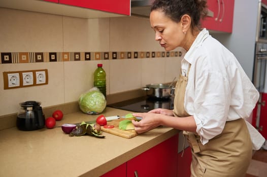 A woman in an apron cutting vegetables in a modern kitchen. Fresh ingredients including lettuce and tomatoes are on the counter, conveying healthy eating and cooking at home.