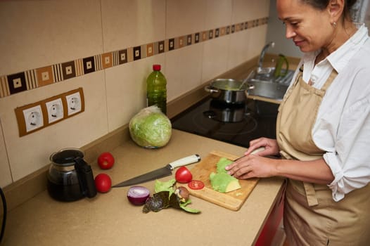A woman in an apron prepares a fresh salad using lettuce, avocado, and tomatoes on a wooden board in a modern kitchen. She embodies healthy living and culinary enjoyment.