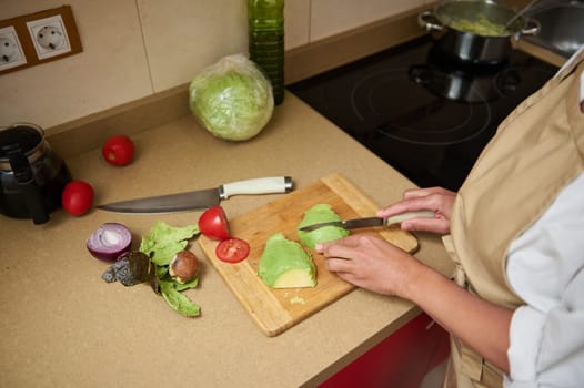 A woman slices avocado on a wooden board in a kitchen with fresh ingredients like lettuce, tomato, and onion, emphasizing healthy meal preparation.