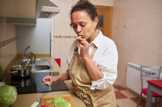 A woman in an apron tastes fresh vegetables in a cozy home kitchen, enjoying the cooking process.