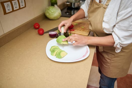 A woman wearing an apron slices avocado in a cozy kitchen setting. Fresh vegetables like tomatoes and onions surround her, creating a warm, healthy, and inviting atmosphere.