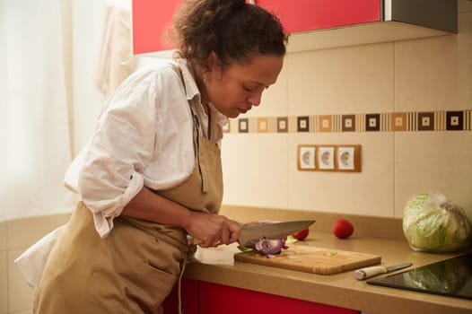 A woman in a white shirt and apron is chopping vegetables on a cutting board in a modern kitchen, showcasing dedication to cooking and healthy lifestyle. The kitchen is bright and functional.