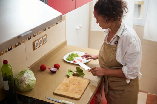 A woman in an apron chops vegetables for a salad in a kitchen. Bright vegetables and fresh greens add a healthy and vibrant touch to the scene.