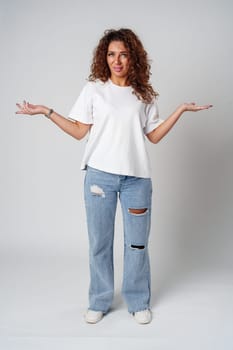 Cheerful woman with curly hair poses with open arms in a white shirt against a plain background close up