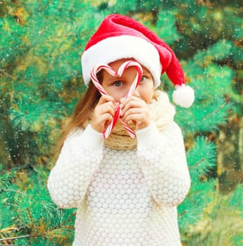Christmas child little girl in santa red hat playing with sweet lollipop cane near a tree