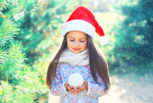 Christmas child little girl in santa red hat holding snowball near a branch tree