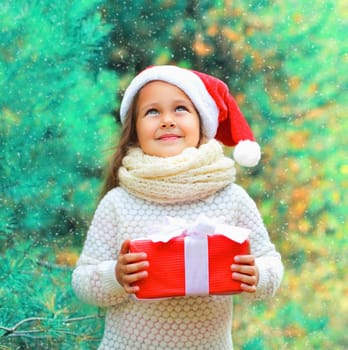 Christmas child little girl in santa red hat with gift box dreaming near a tree