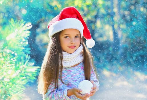 Christmas child little girl in santa red hat with snowball near a branch tree