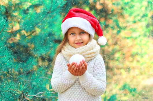 Christmas portrait smiling child little girl in santa red hat holding snowball near tree