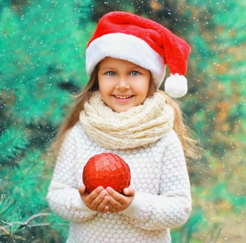 Christmas smiling child little girl in santa red hat with ball near a tree