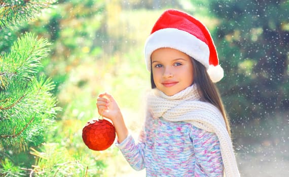Christmas smiling child little girl in santa red hat with ball near a branch tree