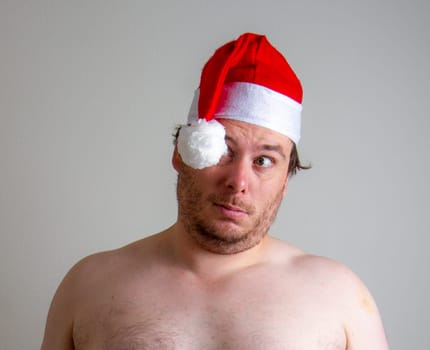 A shirtless man wearing a red Santa hat looks confused against a plain backdrop, capturing a humorous and casual holiday vibe.