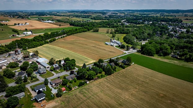 This aerial perspective showcases sprawling farmland, featuring fields of varying colors, houses lined along a road, and gently rolling hills in the background under a sunny sky.