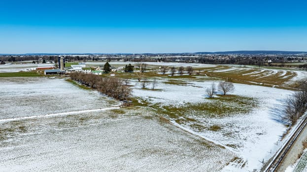 The expansive farm field is covered in a light dusting of snow, showcasing a picturesque winter landscape under a bright blue sky. Farm buildings and trees line the horizon.