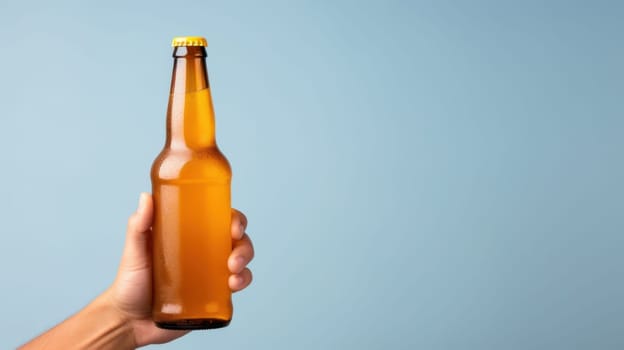 A hand holds a glass beer bottle with a yellow cap against a blue background. The transparent bottle reveals the beer inside, showcasing this popular alcoholic beverage made from barley.