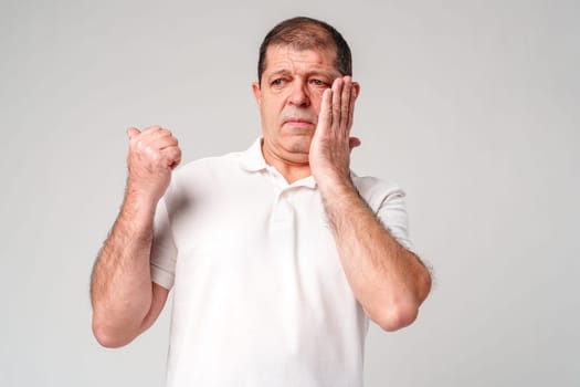 An older man holds his cheek, showing concern, against a plain background close up