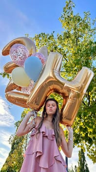 Smiling caucasian young girl model holding bunch of golden balloons smiling at camera. Blonde girl is wearing silk dress against blue sky. Birthday and celebration concept. Vertical