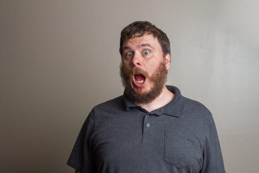 A bearded man shows surprise and shock with wide eyes and open mouth against a neutral background, capturing an intense emotion. Ideal for depicting astonishment or unexpected situations.
