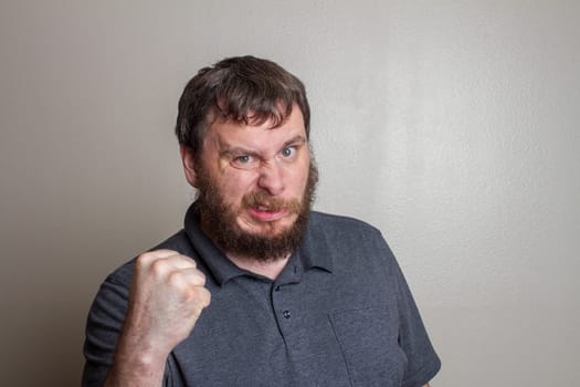 A bearded man expressing anger and frustration with a clenched fist. The intense facial expression captures a moment of strong emotion in a neutral background.