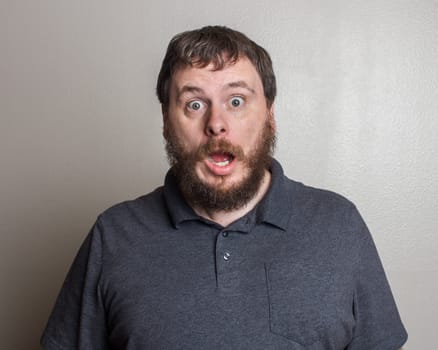 A bearded man with a surprised expression stands against a plain background, conveying shock and amazement. His open mouth and wide eyes capture an unexpected moment.