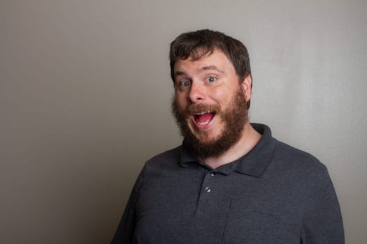 A joyful bearded man with an enthusiastic expression, wearing a grey shirt against a plain backdrop. Captures a moment of happiness and surprise, showcasing an exuberant personality and cheerful mood.