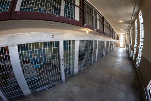 A desolate prison hallway lined with metal jail cells, highlighting the stark and isolating atmosphere of incarceration. The corridor is illuminated by natural light through barred windows.