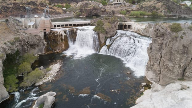 A stunning waterfall cascades over rugged cliffs next to a power plant, set against a scenic landscape. The powerful flow creates a mesmerizing natural scene from shoshone falls in twin falls idaho