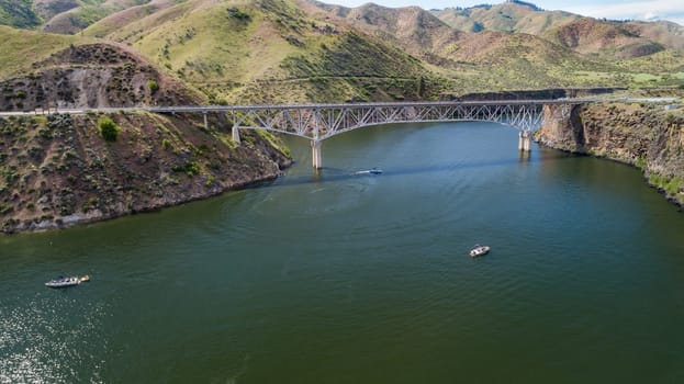 A stunning aerial shot captures a beautiful bridge stretching across a tranquil river gorge, surrounded by lush green hills and occasional boats gliding on the water.