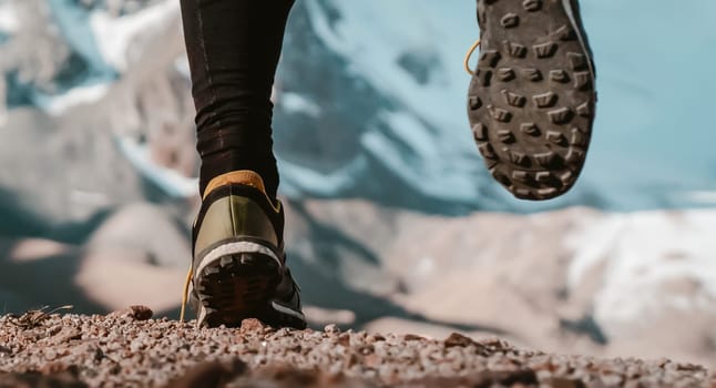 Athlete runner legs run on a trail along a rocky path close-up. Fitness and warm-up, young man with snow-covered mountain peaks in the background and open space around him at sunset.