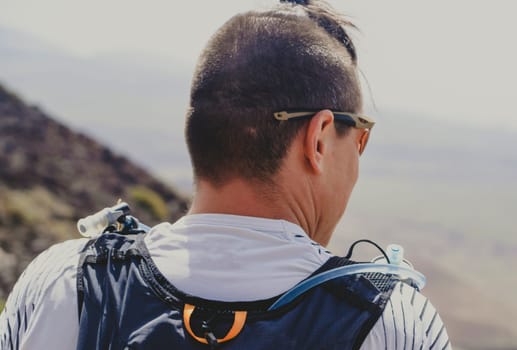 Young athlete with backpack and hydration system for cross-country running preparing for marathon, mountain run on sunny day. Runner drinks water while jogging outdoors, enjoying beautiful landscapes.