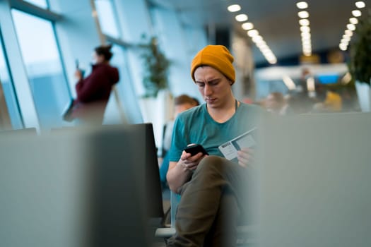A young man sits at the airport and checks in for the next flight via an app on his phone, waits for the plane and works while traveling.