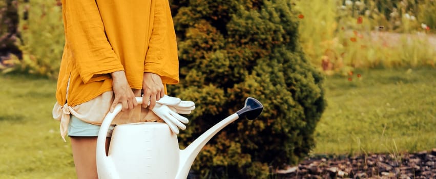 A young girl with gloves and rubber boots is going to water plants in her beautiful summer garden. A professional woman gardener with a watering can is irrigating her lawn and flowers.