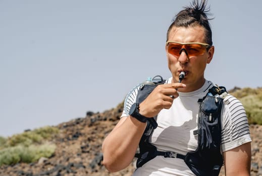 Young athlete with backpack and hydration system for cross-country running preparing for marathon, mountain run on sunny day. Runner drinks water while jogging outdoors, enjoying beautiful landscapes.