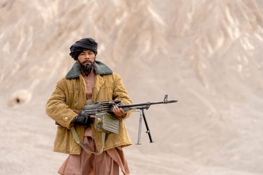 The man with costume look like terrorist hold gun and stand in front of mountain and look at camera.