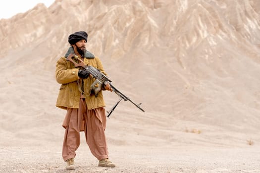 The man with costume look like terrorist hold gun and stand in front of mountain and look forward.