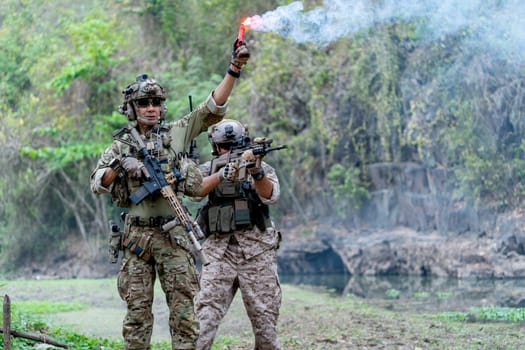 Two militaries or soldiers stay in battlefield near the river with one hold flare fireworks and one stand with point gun forward direction during outdoor practice.