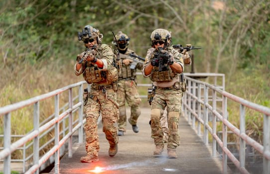 Group of military or soldier team with battle suit walk on the bridge near flare firework and point gun in forward direction during outdoor practice.