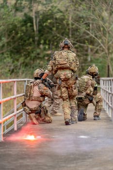 Vertical image back of of military or soldier team with battle suit stay on the bridge near flare firework in position for battle and point gun different direction during outdoor practice.