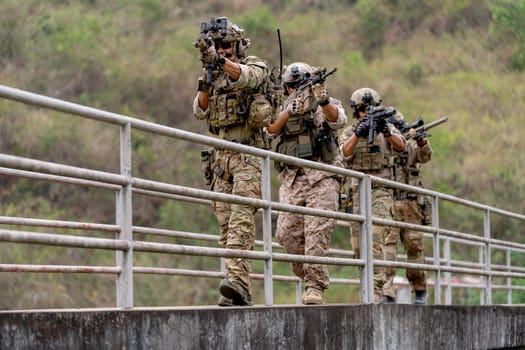 Group of militaries or soldiers with battle suit hold and point gun and walk in the row on bridge during outdoor practice.