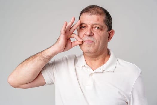 A man makes a playful gesture with his hand near his mouth while smiling in a neutral backdrop close up