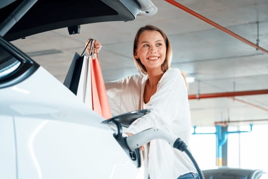 Young woman travel with EV electric car to shopping center parking lot charging in downtown city showing urban sustainability lifestyle by green clean rechargeable energy of electric vehicle innards