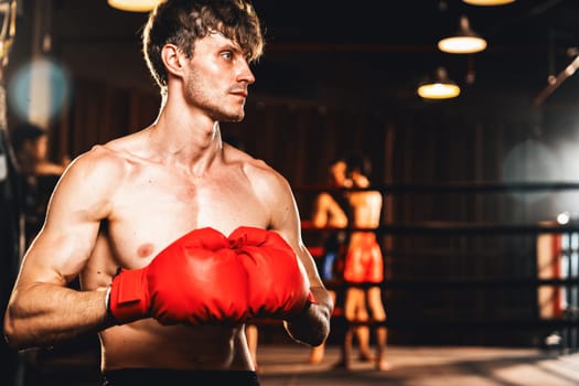 Boxing fighter posing, caucasian boxer put his hand or fist wearing glove together in front of camera in aggressive stance and ready to fight at gym with kicking bag and boxing equipment. Impetus