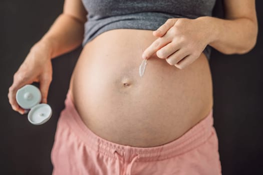 Positive young pregnant woman in comfortable homewear applying belly butter on her big tummy, woman belly.