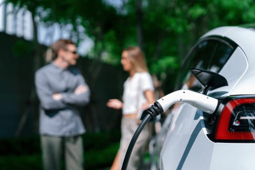 Young couple travel with EV electric car charging in green sustainable city outdoor garden in summer shows urban sustainability lifestyle by green clean rechargeable energy of electric vehicle innards