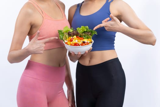 Two young sporty Asian women in sportswear holding salad bowl fill with fruit and vegetable. Natural youthful and fit body lifestyle people with balance nutrition on isolated background. Vigorous