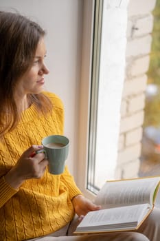 Young beautiful woman sitting by the window yellow knitted sweater read book, daily planner, notepad. Relax concept. Hold cappuccino glass of coffee with white foam. Text is out of focus