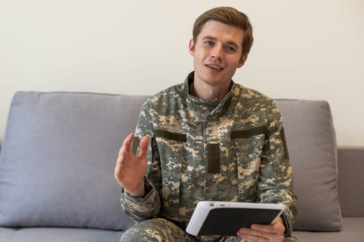 Smiling millennial man in camouflage uniform holding modern digital tablet, chatting with family, posing on white studio background, copy space. Modern technologies and military personnel concept
