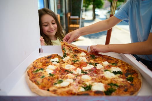 Happy smiling little girl looking at a cardboard box with Neapolitan traditional Italian Margherita pizza, freshly baked with a crispy crust. Italian cuisine, culture and traditions