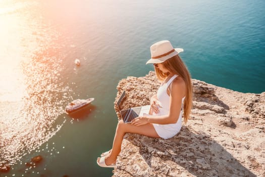 Successful business woman in yellow hat working on laptop by the sea. Pretty lady typing on computer at summer day outdoors. Freelance, travel and holidays concept.